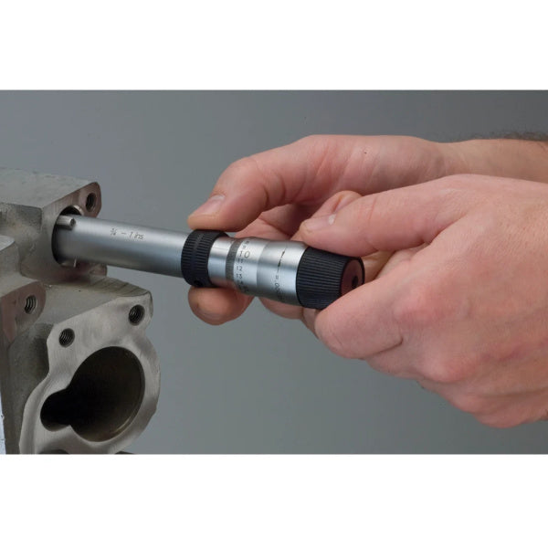 A close-up of a person's hands using a dial indicator to measure a metal part against a plain gray background.